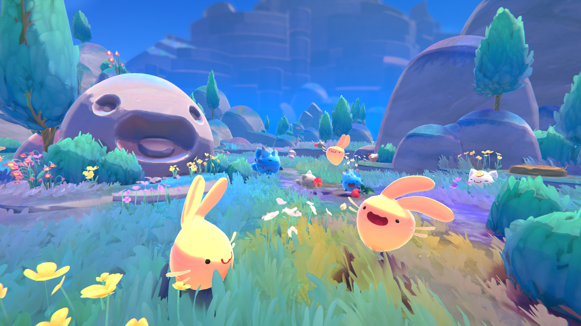 A screenshot from Slime Rancher. There is a grassy field with 
	flowers in the foreground and rocky mountains in the background. On the middle left is a smiling slime boulder. 
	Cone-like trees are dispersed throughout the scenery. There are slimes visible in the image, the most prominent 
	being two happy bunny slimes. The scenery is rich with color.